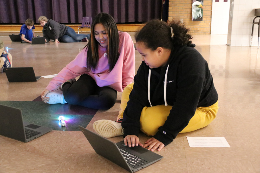 two students work on computers while watching their Ozobots